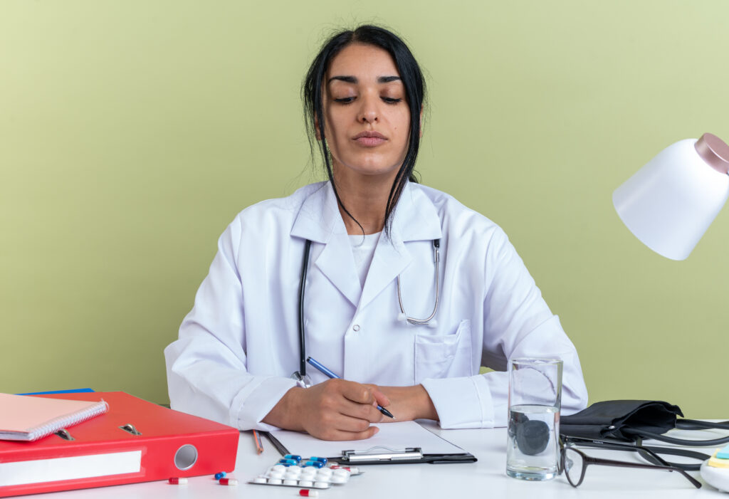 The current image has no alternative text. The file name is: confident-young-female-doctor-wearing-medical-robe-with-stethoscope-sits-desk-with-medical-tools-writing-something-clipboard-isolated-olive-green-wall-scaled.jpg