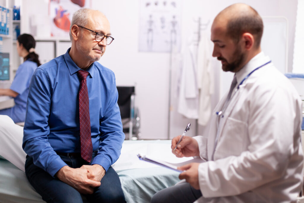 The current image has no alternative text. The file name is: surgeon-wearing-stethoscope-discussing-treatment-examination-room-with-senior-man-1-scaled.jpg